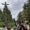 A group of people stand outdoors around a guide near tall totem poles, with trees and a cloudy sky in the background.