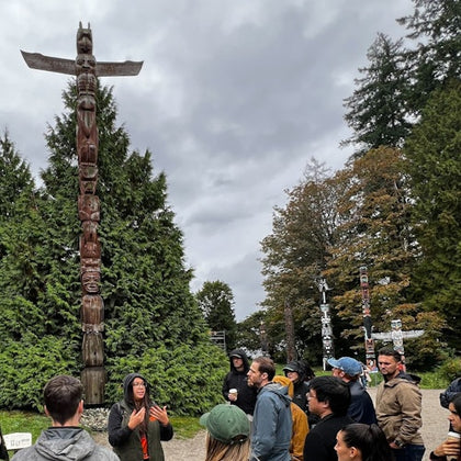 A group of people stand outdoors around a guide near tall totem poles, with trees and a cloudy sky in the background.