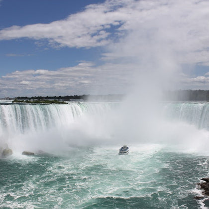 A boat approaching a large, misty waterfall under a partly cloudy sky, surrounded by green water and rocky edges.