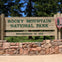 Sign for Rocky Mountain National Park, established in 1915, made of wood and stone, with trees in the background.