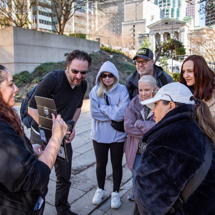A group of six people gathered outdoors, attentively looking at an object held by a woman. Urban buildings are in the background.