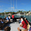 A group of people seated on an open boat cruising in a marina with palm trees and luxury yachts in the background.