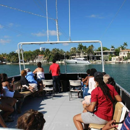 A group of people seated on an open boat cruising in a marina with palm trees and luxury yachts in the background.