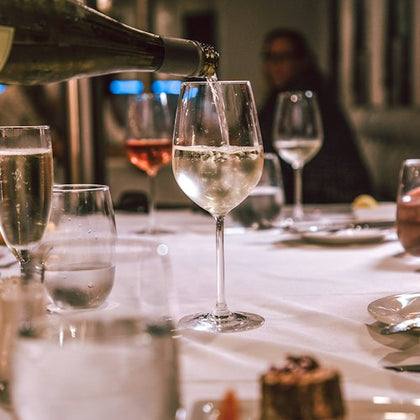 Wine being poured into a glass on a dining table set with glasses, plates, and a dessert in the foreground.