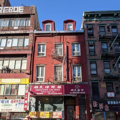 A red brick building with businesses on the ground floor, surrounded by similar multi-story buildings in an urban area.