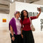 Two women smiling and taking a selfie in a modern art gallery, with white walls and colorful artwork in the background.