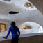 A person in a blue shirt stands inside a multi-level building with curved, irregularly shaped openings. Visitors observe from upper levels.