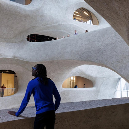 A person in a blue shirt stands inside a multi-level building with curved, irregularly shaped openings. Visitors observe from upper levels.