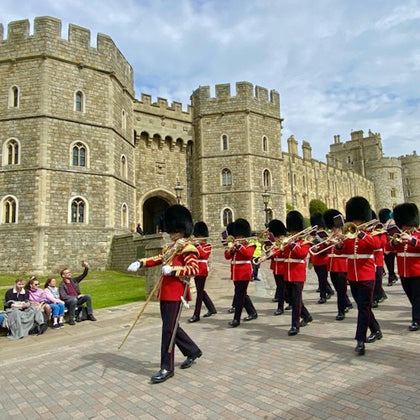 A marching band in red uniforms and black hats parades outside an old stone building, while spectators watch from a bench.