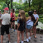 A group of people standing on a pathway in a park, surrounded by trees and greenery. Some are wearing casual summer clothing.
