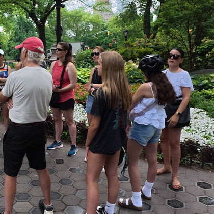 A group of people standing on a pathway in a park, surrounded by trees and greenery. Some are wearing casual summer clothing.
