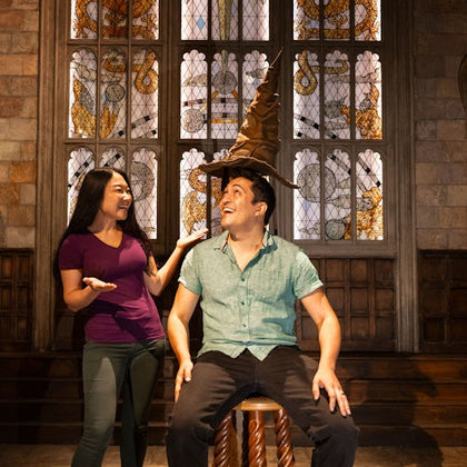 Two people interacting in a room with stained glass windows; one seated, wearing a hat resembling a large wizard's hat, and smiling.