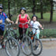 Three people wearing helmets, holding bicycles, and standing on a park path with trees and grass in the background.