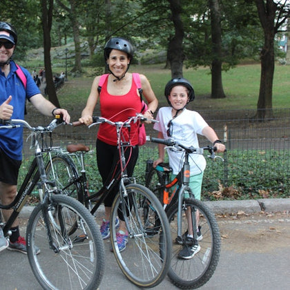 Three people wearing helmets, holding bicycles, and standing on a park path with trees and grass in the background.
