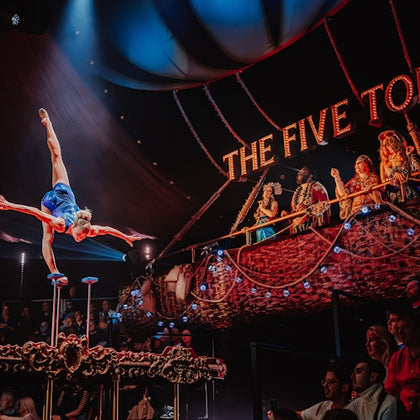 An acrobat performs on hand-balancing props in a circus tent while spectators watch from ornate seating labeled "The Five Toes."