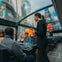 Four people sit at a table inside a glass-roofed bus, one woman standing, with Times Square billboards visible outside.