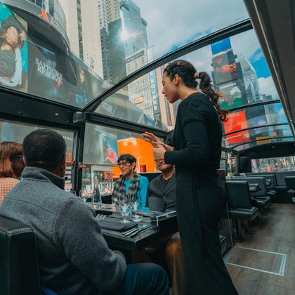 Four people sit at a table inside a glass-roofed bus, one woman standing, with Times Square billboards visible outside.