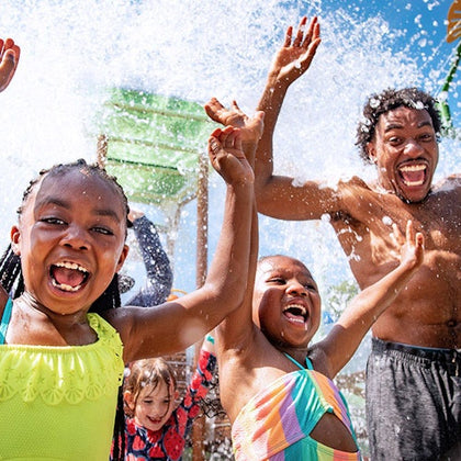 Three children and one man, all in swimsuits, joyfully raise their arms as water splashes over them at a water park.