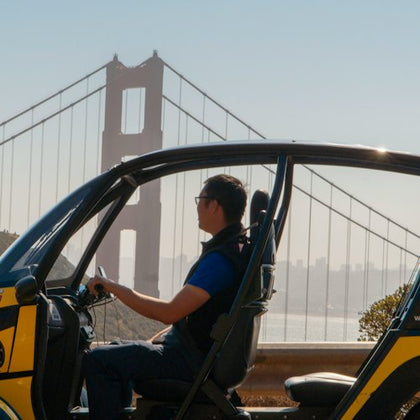 A person in a small enclosed vehicle with the Golden Gate Bridge visible in the background on a sunny day.