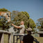 A person feeds a giraffe over a wooden fence at a zoo, with another person observing in the foreground, under a clear sky.