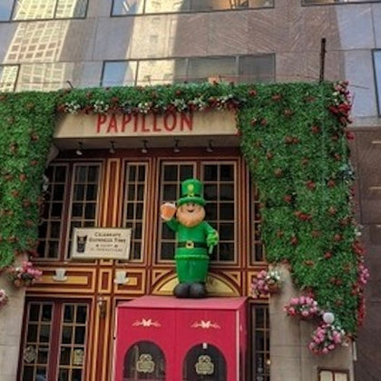 A building entrance decorated with green foliage, flowers, and a cheerful leprechaun figure, with the sign 'PAPILLON' above.