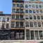 A city street with historic-style buildings, featuring fire escapes and storefronts, under a clear blue sky.