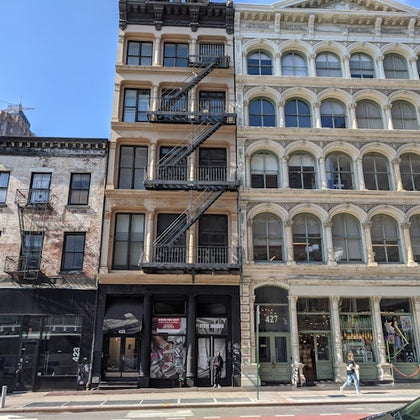 A city street with historic-style buildings, featuring fire escapes and storefronts, under a clear blue sky.