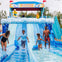 A family enjoys a water slide attraction, with a colorful starting gate and splash pools, surrounded by a sunny outdoor setting.