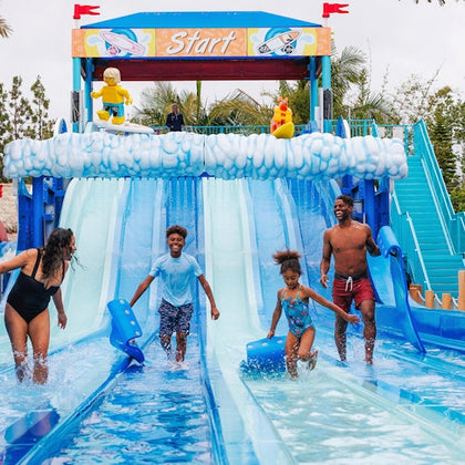 A family enjoys a water slide attraction, with a colorful starting gate and splash pools, surrounded by a sunny outdoor setting.