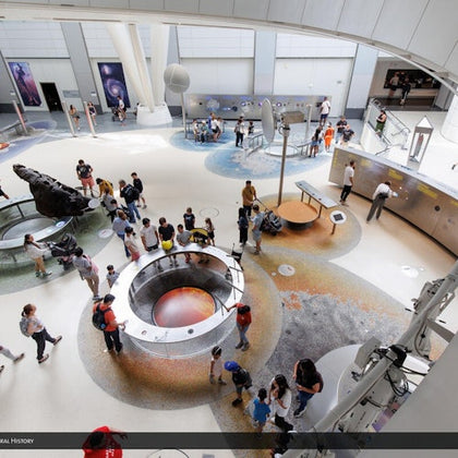 A museum gallery with visitors exploring interactive exhibits and displays, including a circular pit with a glowing center.