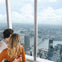 A couple embraces while looking out from a high observation deck window, overlooking a sprawling cityscape with rivers and skyscrapers.