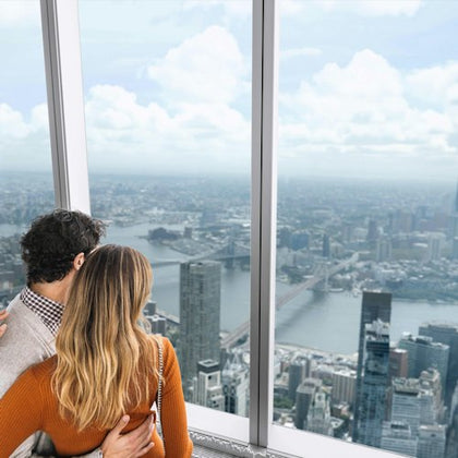 A couple embraces while looking out from a high observation deck window, overlooking a sprawling cityscape with rivers and skyscrapers.