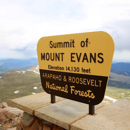 A sign for Mount Evans summit at 14,130 feet, located in the Arapaho and Roosevelt National Forests, with scenic views.