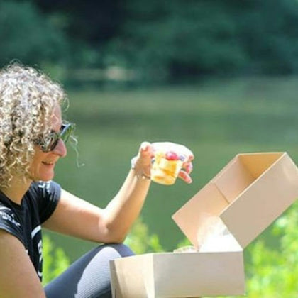 A person with curly hair and sunglasses sits outdoors, holding a drink near an open cardboard box, with greenery and water in the background.