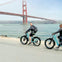 Two people riding bicycles on a waterfront path with the Golden Gate Bridge in the background on a cloudy day.