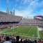 A football stadium filled with spectators, players on the field, and a city skyline in the background under a blue sky.