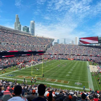 A football stadium filled with spectators, players on the field, and a city skyline in the background under a blue sky.