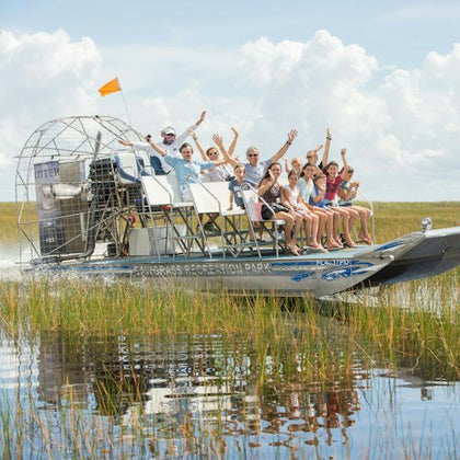 A group of people riding an airboat in a grassy wetland area under a cloudy sky, waving and smiling enthusiastically.