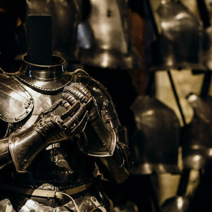 A suit of medieval armor with clasped gauntlets on display, surrounded by other armors in the background.