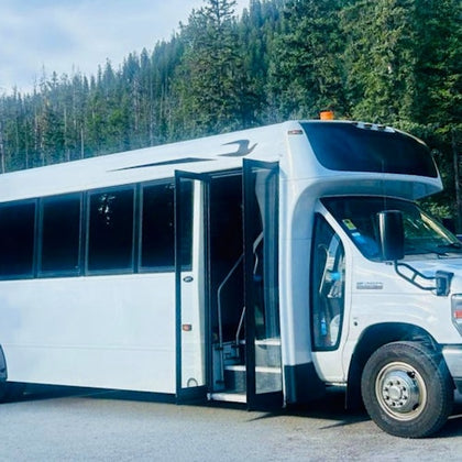 White shuttle bus with open doors parked on a road surrounded by dense evergreen trees under a cloudy sky.