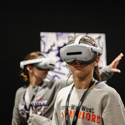 Two individuals wearing VR headsets in a dimly lit environment, one in the foreground and one slightly blurred in the background.