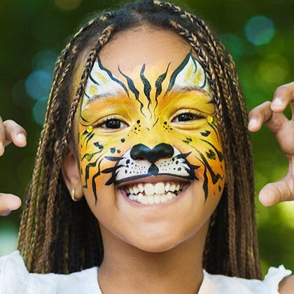 Child with face painted like a tiger, smiling and posing with hands raised, against a blurred green background.