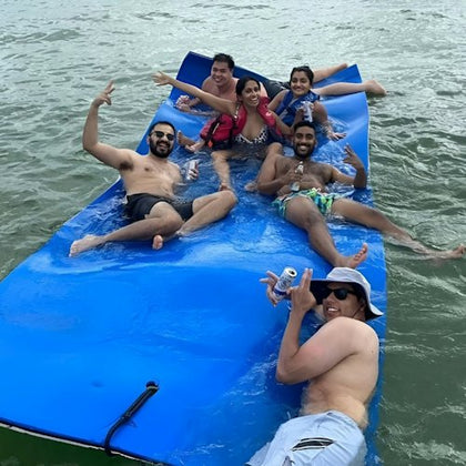 Six people relaxing on a blue floating mat in the water, holding drinks and making gestures, enjoying a sunny day.