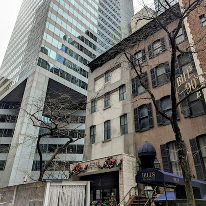 A street-level view of a modern high-rise next to older townhouses, one of which has a canopy labeled "Bill's."