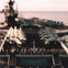 Aircraft carrier deck with several fighter jets lined up, surrounded by sailors standing at attention along the edges.