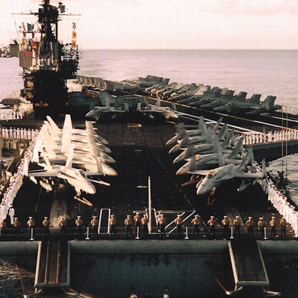 Aircraft carrier deck with several fighter jets lined up, surrounded by sailors standing at attention along the edges.