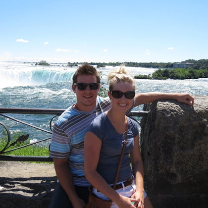 Two people wearing sunglasses posing near a stone railing with a large waterfall and greenery in the background.