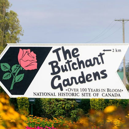 Sign reading "The Butchart Gardens" with a pink rose illustration, surrounded by flowers and greenery, under a clear sky.
