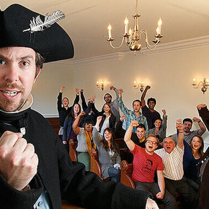 A man in historical attire gestures in the foreground while a group of people behind him cheer enthusiastically indoors.