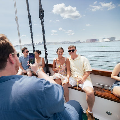 Three people pose for a photo on a boat while another person takes the picture. Water and city skyline are in the background.